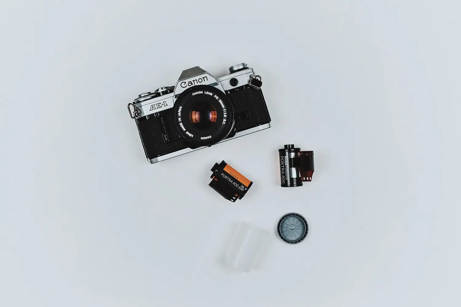 A film camera, rolls of film and a lens cap laid out on a white surface.