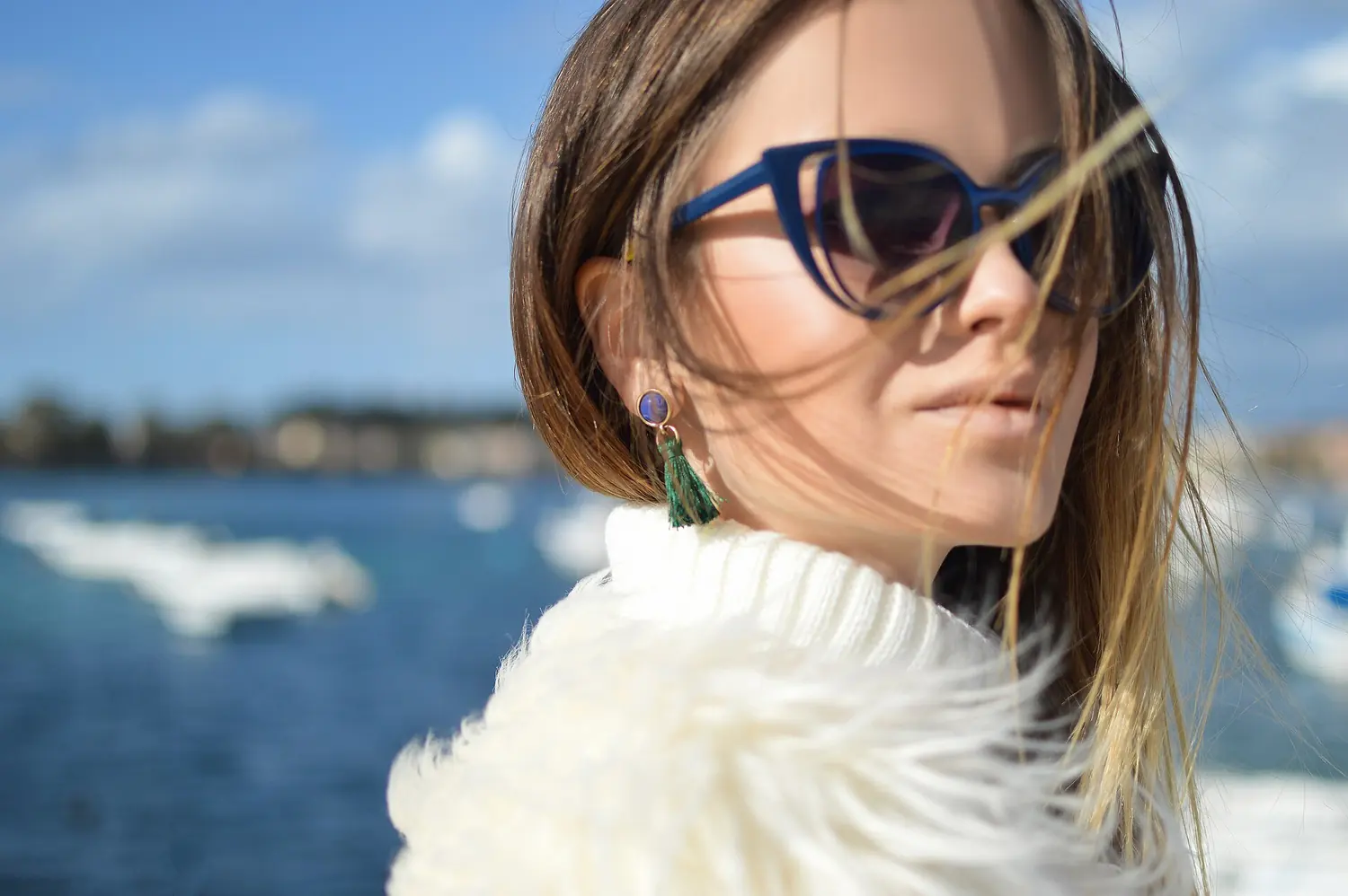Woman in blue sunglasses and a white faux fur coat by the sea.
