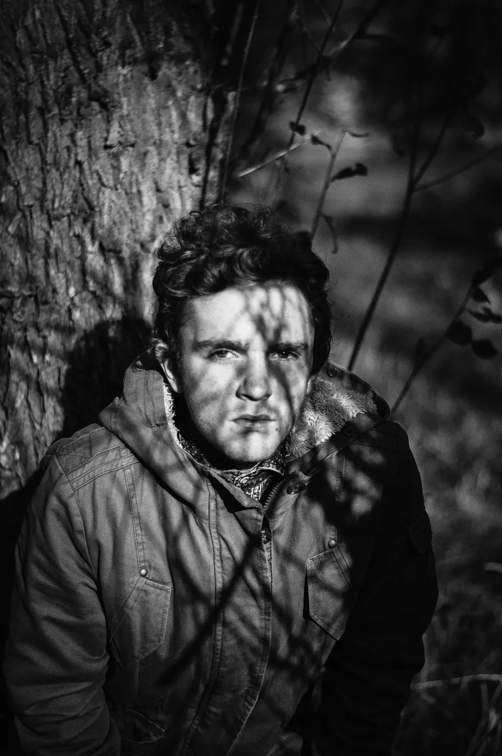 Black and white portrait of a man in dappled shade against a tree.