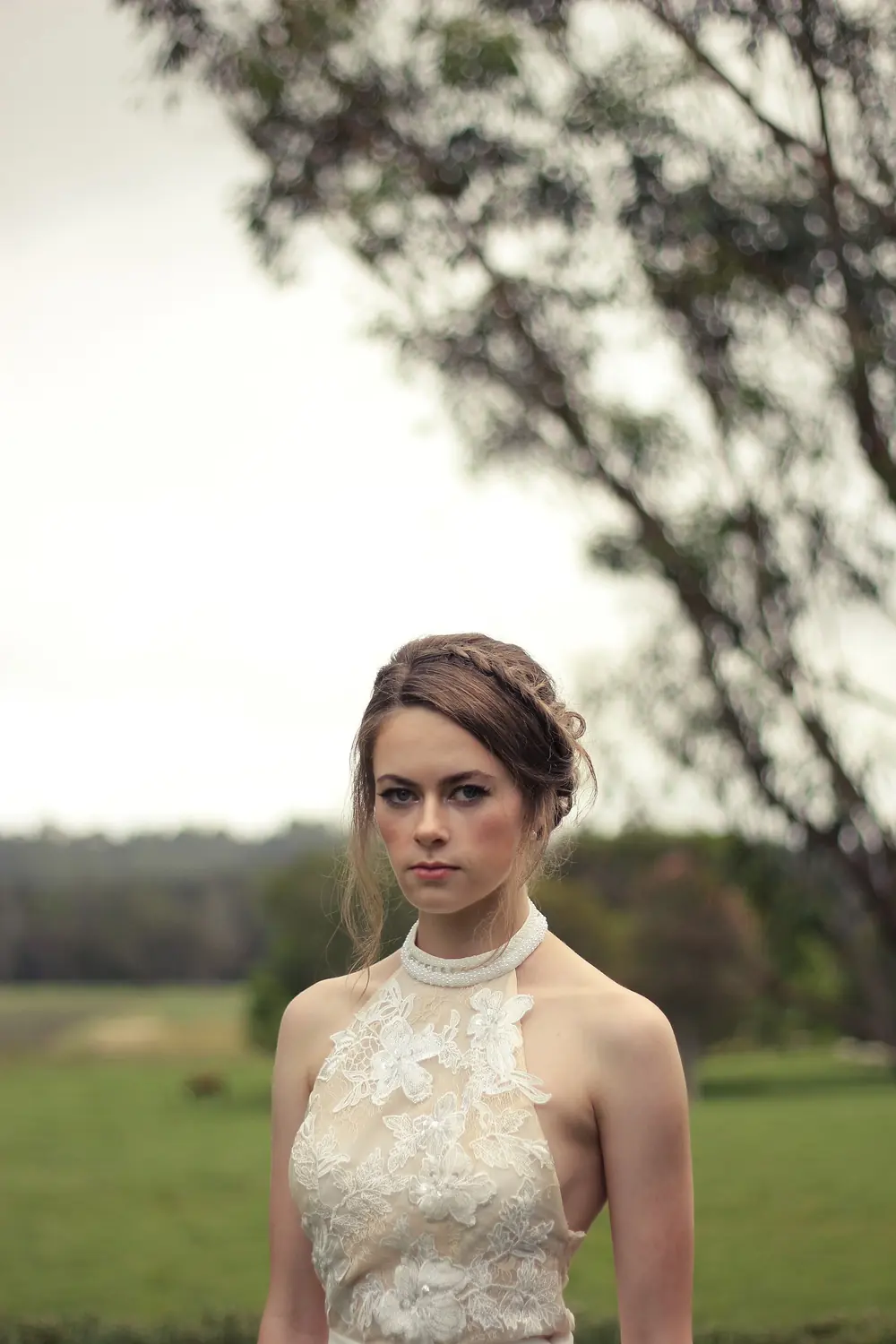 Bride in a lace dress standing in a field at the edge of a wood.
